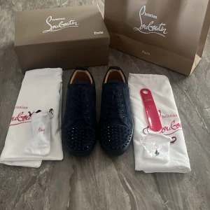 Mörkblåa sneakers med nitar från Christian Louboutin - Säljer ett par mörkblåa sneakers från Christian Louboutin med ikoniska röda sulor och svarta nitar på tån. Skorna har snörning och är tillverkade i skinn. Kommer med originalkartong, dustbags och tillbehör.
