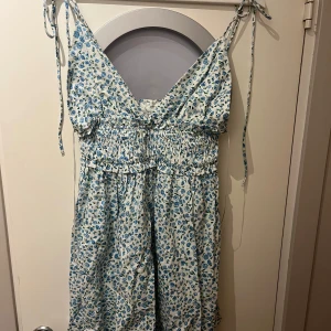 Blommig sommarklänning från Zara - Supersöt ljus klänning från Zara med blått blommönster och smock i midjan. Axelbandslös modell med knytbara axelband och v-ringad hals. Perfekt för både fest och vardag. Säljer då jag tycker den blir lite stor över bysten på mig. 