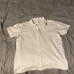 Vit pikétröja från Zara - Säljer en klassisk vit pikétröja från Zara i storlek 140. Tröjan har korta ärmar, krage och knäppning framtill. Perfekt för en clean och stilren look. Tillverkad i bomull för en skön känsla.