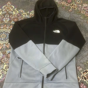 Svart och grå hoodiejacka från The North Face - Säljer en snygg hoodiejacka från The North Face i svart och ljusgrått. Jackan har dragkedja framtill, två sidofickor med dragkedja och en stor huva. Perfekt för lager-på-lager och streetwear.