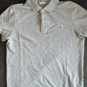 Grå pikétröja från Moncler - Snygg grå pikétröja från Moncler med klassisk krage och tre knappar framtill. Tröjan har Moncler-logga broderad på bröstet och är tillverkad i bomull. Perfekt för en stilren och avslappnad look.