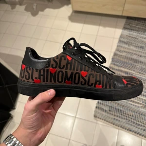 Svarta sneakers från Moschino med röda hjärtan - Säljer ett par svarta sneakers från Moschino med stora logotexter och röda hjärtan över hela skon. Skorna har svarta snören och en svart sula med Moschino-logga undertill. Perfekt statement-piece för dig som gillar att sticka ut.