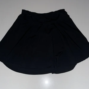 Svart träningskjol från BOW19 - Säljer en svart träningskjol från BOW19 med inbyggda shorts under. Kjolen har en sportig look och elastisk midja för extra komfort. Perfekt för dig som gillar att röra på dig och vill ha en stilren och enkel kjol till träningen.