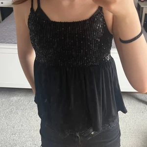Svart glittrig linnetopp - Säljer en svart linnetopp med smala axelband och glittriga paljettdetaljer upptill. Toppen har en luftig passform nedtill och är perfekt för dig som vill sticka ut lite extra.