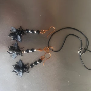 Svart bi-smyckesset med örhängen och halsband - Unikt smyckesset med svarta bi-örhängen och matchande halsband. Örhängena har svarta pärlor och detaljer i guld, och bina är detaljerade med vingar och kropp. Halsbandet har svart snöre och silverfärgat lås.