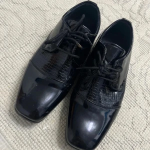 Svarta lackade snörskor med fyrkantig tå - Säljer ett par svarta snörskor i lack med fyrkantig tå och låg klack. Skorna har ett stilrent mönster på ovandelen som ger en extra touch. Perfekta för dig som vill sticka ut med en klassisk men modern look. För barn.