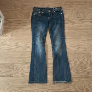 Blå Miss Me jeans med broderade bakfickor - Säljer ett par blå jeans från Miss Me med snygga broderade detaljer och nitar på bakfickorna. Jeansen har bootcut-modell och klassisk femficksdesign. Märkeslogga på bakre linningen och slitningar framtill för en cool look.