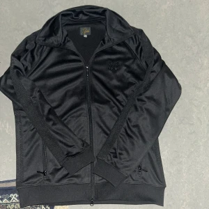 Svart track jacket från Needles - Säljer en svart track jacket från Needles med dragkedja och broderad fjäril på bröstet. Jackan har långa ärmar, ribbade muddar och är tillverkad i glansig polyester. Klassisk sportig look med diskreta detaljer längs ärmarna. Aldrig använd, storlek S. Tröjan var även köpt i Japan. Pris kan diskuteras.
