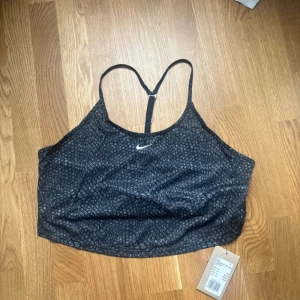 Svart mönstrad linnetopp från Nike - Säljer en snygg svart linnetopp från Nike med ett diskret mönster och vit logga framtill. Toppen har smala axelband och en croppad passform, perfekt för träning eller en avslappnad stil.