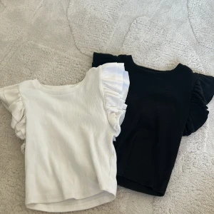 Svart och vit ribbad topp med volangärmar - Säljer två ribbade toppar i svart och vitt med volangdetaljer på ärmarna. Perfekt för en stilren look med en twist. Topparna är ärmlösa och har en bekväm passform.