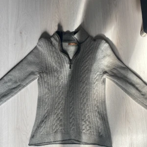 Grå stickad tröja från Jerem - Säljer en stilren grå stickad tröja från Jerem. Tröjan har en dragkedja vid halsen och ett snyggt flätmönster på framsidan. Den är tillverkad i en mjuk blandning av lammull och kashmir, vilket gör den både varm och bekväm. Perfekt för kyliga dagar! (Ps: Storlek S men sitter som XS)