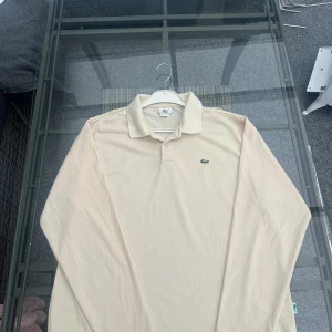 Beige pikétröja från Lacoste - Säljer en klassisk beige pikétröja från Lacoste med den ikoniska gröna krokodilloggan broderad på bröstet. Tröjan har krage, knappar vid halsen och är långärmad. Perfekt för en stilren och avslappnad look.