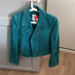 Turkos mockajacka från Juna - Snygg turkos mockajacka från Juna i bikerstil. Jackan har en asymmetrisk dragkedja och två fickor med dragkedjor. Perfekt för att ge din outfit en cool touch ,  lapp kvar säljer då den inte används 