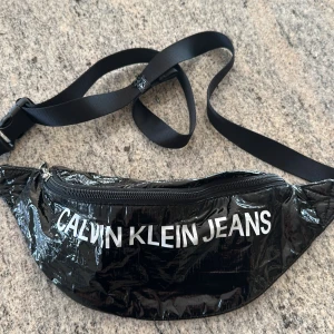 Svart magväska från Calvin Klein Jeans - Snygg svart magväska från Calvin Klein Jeans i glansigt syntetmaterial. Väskan har justerbart band, dragkedja upptill och logga både fram och bak. Perfekt för att bära det viktigaste med stil.