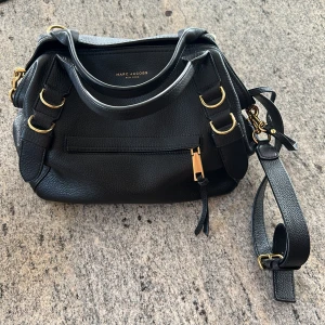 Svart handväska från Marc Jacobs - Snygg svart handväska i skinn från Marc Jacobs med guldfärgade metalldetaljer och justerbar axelrem. Väskan har dragkedja framtill, innerficka med dragkedja och medföljande liten plånbok. Perfekt för dig som vill ha en stilren accessoar. Man får 5% RABATT när följer NordqvistFashionCloset på Instagram samt här på plick!!😊Mvh Wilgot