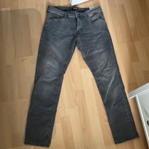 Grå jeans från Jack & Jones - Säljer ett par grå jeans från Jack & Jones med klassisk femficksdesign och raka ben. Jeansen har dragkedja och knapp i gylfen samt diskreta sömmar på bakfickorna. Perfekta för en stilren vardagslook. 