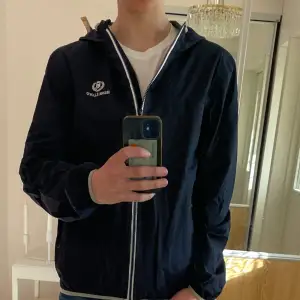 Säljer en mörkblå windbreaker från Henri Lloyd med huva och vit dragkedja. Jackan har logga på bröstet och två sidofickor. Perfekt för vår och sommar.
