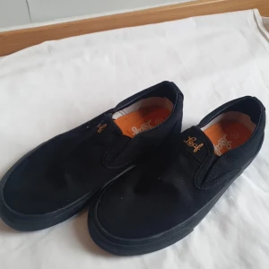 Svarta slip-on sneakers från Leaf - Svarta slip-on sneakers från Leaf med diskret logga på hälen och insidan. Skorna har en platt, orange sula med mönster för bra grepp. Ovandelen är i textil och designen är enkel och stilren, perfekt för en avslappnad look.