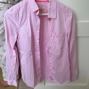 Ljusrosa skjorta från Holly & Whyte - Säljer en klassisk ljusrosa skjorta från Holly & Whyte. Skjortan har lång ärm, bröstficka och knappar framtill. Perfekt för en stilren och enkel look.