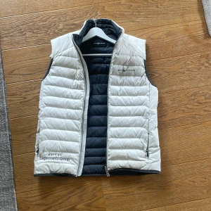 Vit dunväst från Sail Racing - Säljer en ljusgrå dunväst från Sail Racing med svart insida och dragkedja framtill. Västen har broderad logga på bröstet och är lätt vadderad för extra värme. Perfekt för lager-på-lager under kyliga dagar.