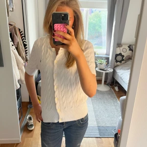 Vit kabelstickad kofta från Polo Ralph Lauren - Säljer en vit kabelstickad kofta från Polo Ralph Lauren med korta ärmar och knappar framtill. Klassisk design med ribbade muddar och liten broderad logga på bröstet. Perfekt till jeans eller kjol!