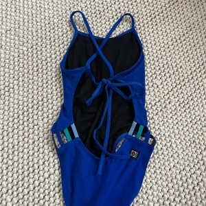 Blå baddräkt från Jolyn med knyt och färgglada band - Säljer en blå baddräkt från Jolyn, modell GAVIN. Baddräkten har smala axelband som korsas i ryggen och knyts, samt färgglada band på sidorna. Storlek 28. Acceptabelt skick. Lite sliten på rumpan, men i övrigt bra skick. 