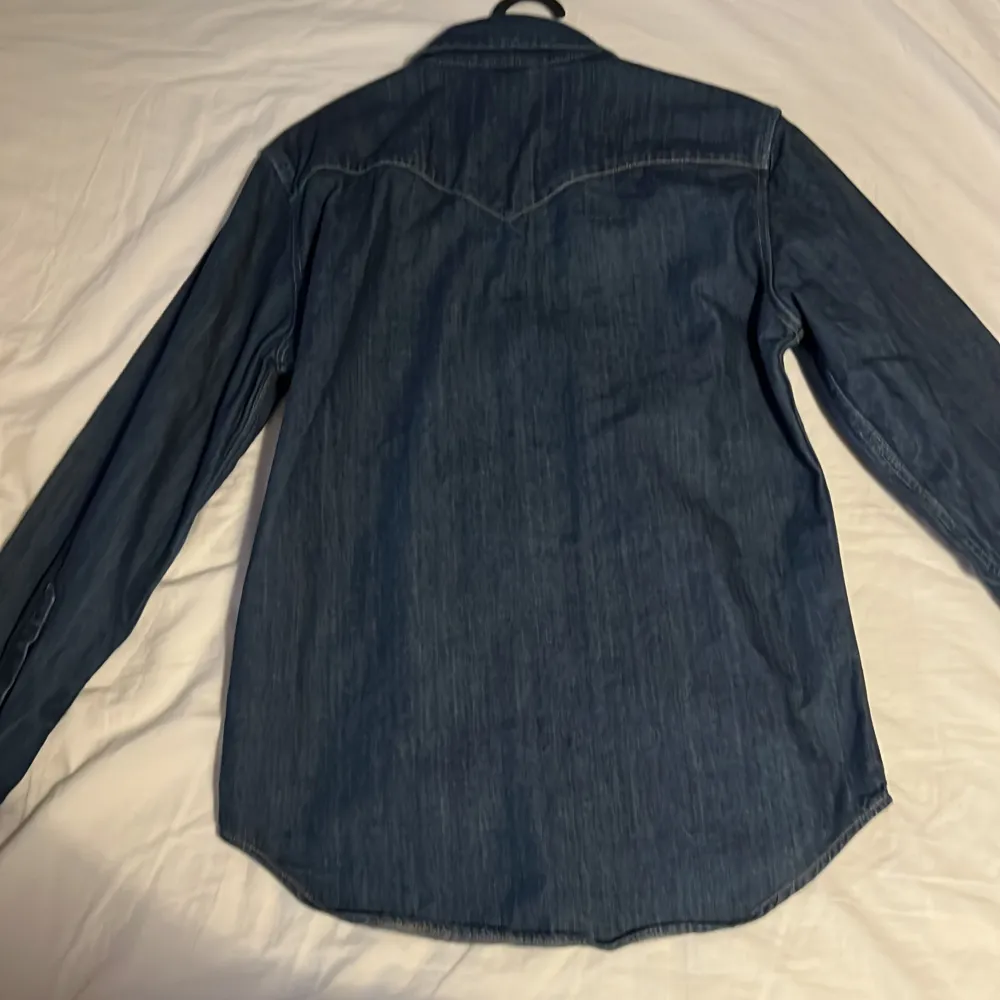 Mörkblå jeansskjorta från Levi's med klassisk westernstil. Skjortan har långa ärmar, tryckknappar och två bröstfickor med lock. Snygga kontrastsömmar och ikonisk röd Levi's-tagg på fickan. Perfekt för en avslappnad look.. Paidat.