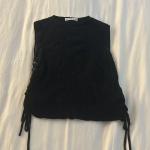 Top med snörning från Zara - Säljer en svart ribbad ärmlös topp från Zara med snörningsdetalj på sidan. Toppen har croppad passform och är tillverkad i stretchigt material. Perfekt att styla med jeans eller kjol.