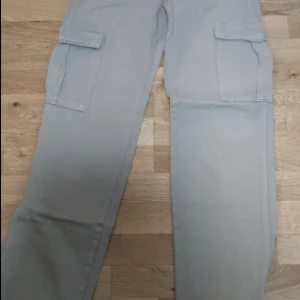 Ljusgrå cargobyxor från Pull&Bear - Säljer ett par ljusgrå cargobyxor från Pull&Bear med stora fickor på sidorna. Byxorna har en rak passform och är perfekta för en avslappnad stil. Tillverkade i ett mjukt bomullsmaterial. Byxorna sälja även på vinted.