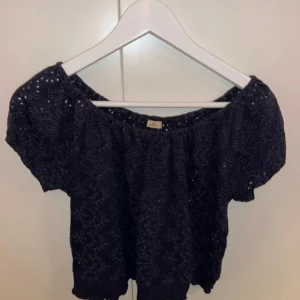 Svart offshoulder blus med broderade detaljer - Säljer en svart offshoulder blus med broderade hålmönster och korta puffärmar. Toppen har en croppad passform och resår vid axlar och midja för en bekväm och snygg look.