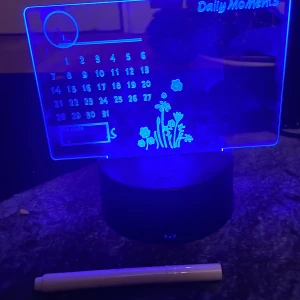 LED-belyst skrivbar kalender 'Daily Moments' - Säljer en transparent akrylkalender med LED-belysning i olika färger. Kalendern har plats för anteckningar, månadsöversikt och dekorativa blommor. Basen är svart och kalendern kan skrivas på med medföljande penna. Perfekt för att hålla koll på viktiga datum.