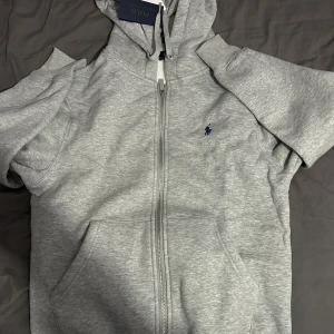 Grå hoodie från Ralph Lauren - Säljer en stilren grå hoodie från Ralph Lauren med dragkedja och en liten broderad logga på bröstet. Perfekt för en avslappnad stil. Den har en klassisk passform och är tillverkad i mjukt material.