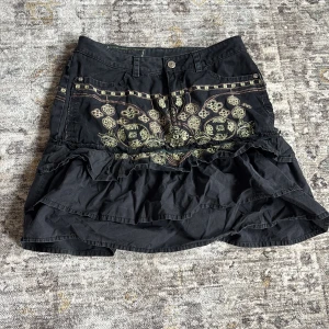 Svart kjol med broderade detaljer från Pepe jeans - Svart kjol från Pepe jeans med broderade mönster i beige och volangdetaljer. Kjolen har är lågmidjad och är kort i modellen. Perfekt för dig som gillar unika plagg med mycket detaljer. 37cm i midjan tvärs över 😇