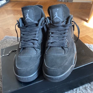Svarta Nike Air Jordans 4 Retro - Säljer ett par helsvarta Nike Air Jordans 4 Retro med klassisk siluett och Jumpman-logga på plösen och hälen. Skorna har snörning, mesh-detaljer och är tillverkade i mocka och syntetmaterial. Perfekta för dig som gillar streetwear och sneakers med attityd.
