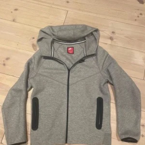 Nike tech grå - Grå hoodiejacka från Nike med svart dragkedja och svarta detaljer vid fickorna. Jackan har huva och lång ärm, perfekt för en sportig stil. Passformen är normal och materialet känns mjukt och bekvämt.