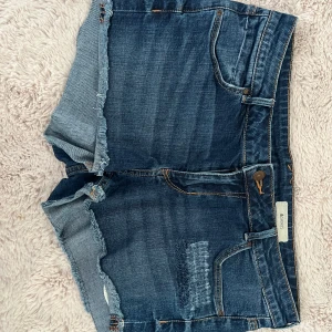 Blå jeansshorts med fransig kant från &Shorts - Snygga blå jeansshorts från &Shorts med fransig kant nertill och slitna detaljer framtill. Klassisk femficksmodell med knapp och dragkedja. Perfekta för varma dagar och enkel att matcha med olika toppar.