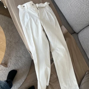 Vita kostymbyxor  - Snygga vita kostymbyxor med hög midja. Byxorna har en klassisk rak passform och är perfekta för en stilren look. De har knappar bak och är tillverkade i ett elegant material.