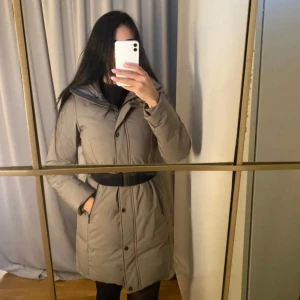 Beige parkajacka med bälte - Snygg beige parkajacka med avtagbart bälte och dragkedja. Jackan har långa ärmar och en stilren design som passar perfekt för kyligare dagar.
