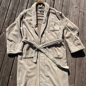 Beige morgon/badrock med broderad figur från Pooh - Säljer en mysig beige morgonrock från Pooh i 100% bomull. Den har en broderad figur på bröstfickan och ett praktiskt bälte i midjan. Perfekt för att hålla sig varm och bekväm hemma. Spår av användning, några fläckar som knappt syns! Skärpet mest nopprigt! Den är i mkt bra skick men blir bara hängande! 