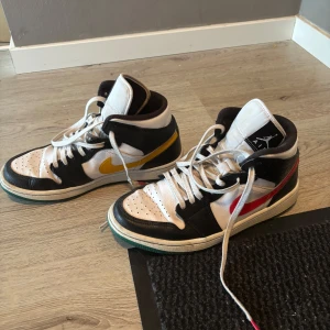 Nike Air Jordans med färgade swooshes - Säljer ett par Nike Air Jordans med svart, vitt och inslag av rött och gult på swooshen. Skorna har högt skaft, snörning och klassisk Jordan-logga på plösen. Perfekta för dig som gillar streetwear och vill sticka ut med färgade detaljer.