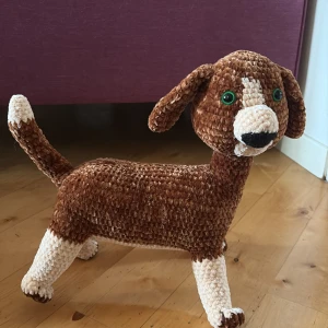 Virkad hund i brunt och beach garn - Säljer en handgjord virkad hund i mjukt brunt och beach garn. Hunden har gröna plastögon, lång svans och hängande öron. Perfekt som gosedjur eller dekoration. Den är ca 30 cm lång och har fina detaljer på nos och tassar.