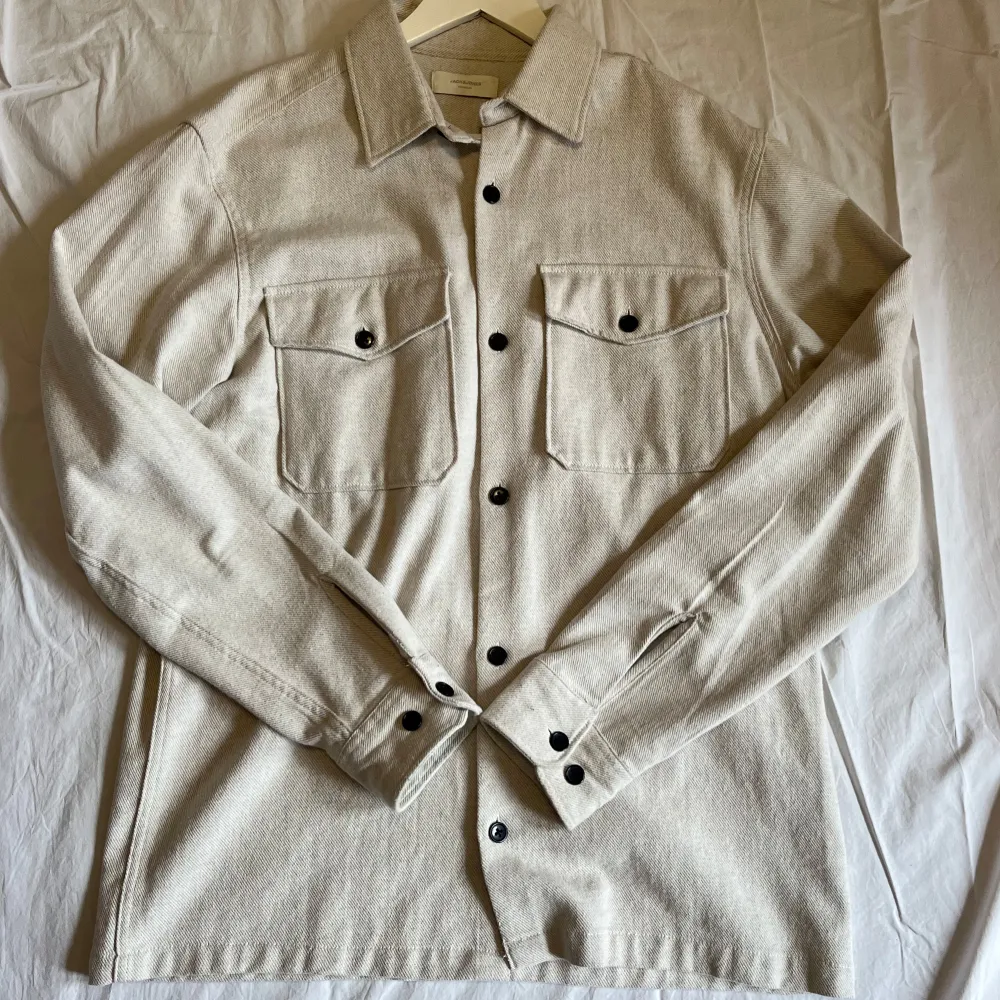 Stilren beige overshirt från Jack and jones med svarta knappar och två stora bröstfickor. Skjortan har klassisk krage och lång ärm med knapp vid manschetten. Perfekt plagg i ett mjukt bomullsmaterial som passar till allt. Takit.