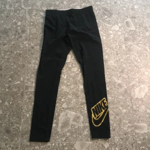 Svarta leggings från Nike med gul logga - Svarta leggings från Nike med stor gul Nike-logga på ena benet. Tight passform och elastisk midja. Perfekta för träning eller en sportig look. Tillverkade i stretchigt material för maximal komfort.