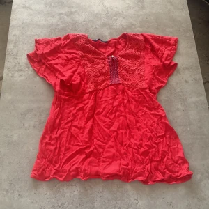 Röd blus från Weekend - Säljer en röd blus från Weekend med vackra spetsdetaljer och en glittrig knapp framtill. Blusen har korta ärmar och ett broderat mönster i blått som ger en fin kontrast. Perfekt för en färgglad look!