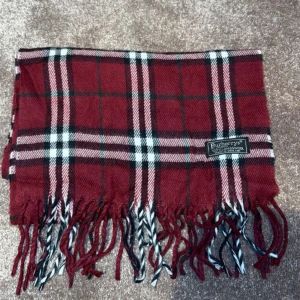  Burberry Halsduk - Snygg rutig halsduk i rött, svart och vitt från Burberry. Den har klassiska fransar i ändarna och ett tidlöst mönster som passar perfekt för kyliga dagar. Perfekt accessoar för att lyfta din outfit!