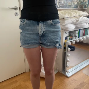 Blå jeansshorts med hål - Säljer ett par blå jeansshorts med coola slitningar och fransiga kanter. Perfekta för sommardagar och ger en avslappnad look. De har en klassisk femficksdesign och är lågmidjade för en bekväm passform.