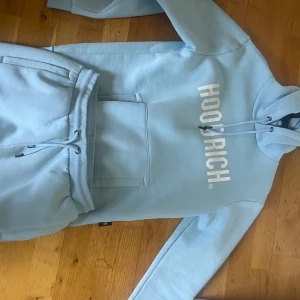 Ljusblå dress från Hoodrich - Säljer en snygg ljusblå dress från Hoodrich med vit text på bröstet. Den har en stor ficka framtill och justerbar huva med dragsko. Perfekt för en avslappnad stil.