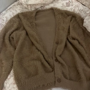 Brun fluffig kofta - Mysig brun kofta med fluffig textur och knappar framtill. Perfekt för kyliga dagar när du vill ha något bekvämt och stiligt. Långärmad design med ribbade muddar vid ärmslut och nederkant.