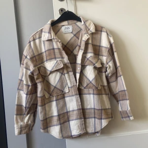 Rutig skjorta/jacka från Zara - Säljer en snygg rutig skjorta från Zara i beige och blå toner. Skjortan har långa ärmar och knappar framtill samt två bröstfickor med lock. Perfekt för våriga kvällar