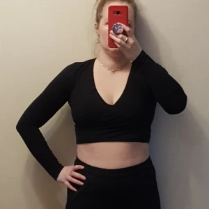 Svart croppad topp från Gina Tricot strl S - Säljer en svart croppad topp från Gina Tricot. Toppen har långa ärmar och en v-ringad design som ger en stilren look. Perfekt för att matcha med höga jeans eller kjol. Topen har v-ringning med omlott look och jag har satt ett litet resårband i urringningen för att den ska sitta på plats den är deför de är rynkigt se bild 3